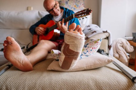 Téléchargez les photos : Homme blessé avec pied bandé et apprendre à jouer de la guitare en ligne. - en image libre de droit