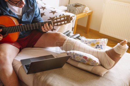 Photo for Injured man with bandaged foot and learn to play guitar online. - Royalty Free Image