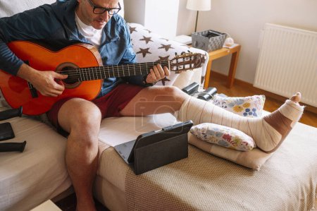 Foto de Hombre lesionado con el pie vendado y aprender a tocar la guitarra en línea. - Imagen libre de derechos