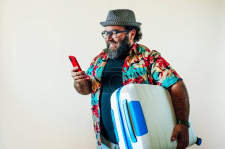 Téléchargez les photos : Homme gras barbu utilisant un téléphone intelligent et une valise de transport pour les voyages. - en image libre de droit