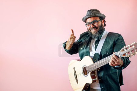 Téléchargez les photos : Heureux homme barbu jouant de la guitare acoustique sur fond rose. Il lève le doigt comme un symbole de possible. - en image libre de droit