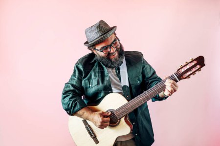Foto de Hombre barbudo feliz tocando la guitarra acústica sobre fondo rosa. - Imagen libre de derechos