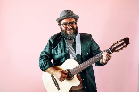 Foto de Hombre barbudo feliz tocando la guitarra acústica sobre fondo rosa. - Imagen libre de derechos