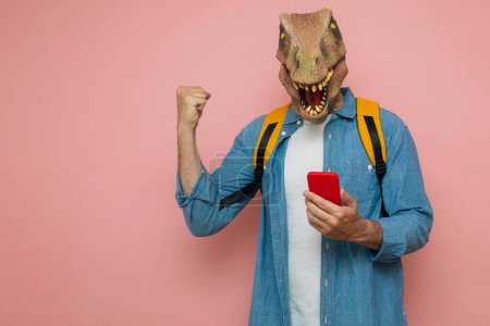 Foto de Hombre eufórico con mochila y máscara de lagarto sobre fondo rosa usando el teléfono. - Imagen libre de derechos