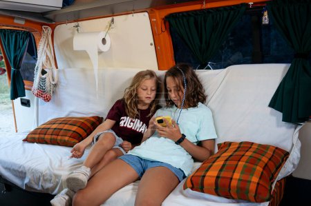 Foto de Dos niños hermanos usando el teléfono inteligente en un maravilloso día acampando en una furgoneta. Concepto Van Life. - Imagen libre de derechos