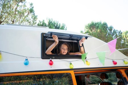 Foto de La chica feliz saca la cabeza por la ventana de la furgoneta en un día maravilloso en el campamento. Concepto Van Life. - Imagen libre de derechos