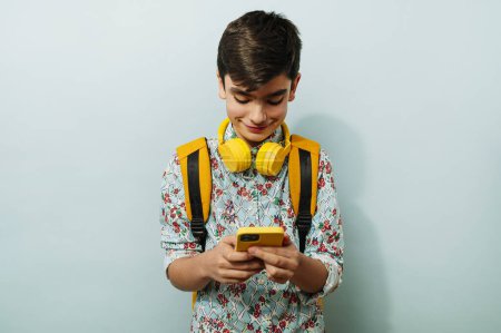 Foto de Chico joven usando teléfonos inteligentes y auriculares amarillos en el estudio sobre fondo azul. - Imagen libre de derechos