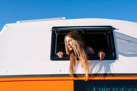 Téléchargez les photos : La fille heureuse sort la tête par la fenêtre du van lors d'une merveilleuse journée au camp. Concept de vie Van. - en image libre de droit