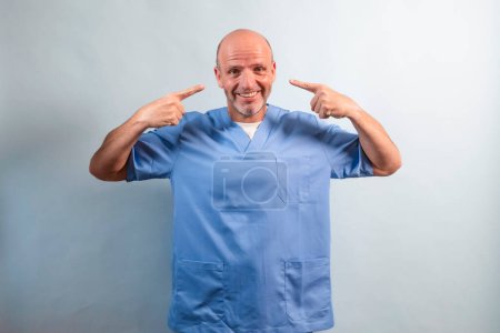 Photo for Portrait of a physiotherapist wearing light blue gown and pointing at his face with his fingers. - Royalty Free Image