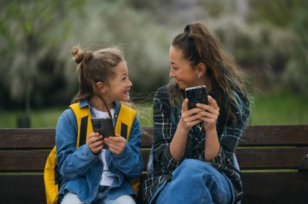 Téléchargez les photos : Petit garçon accompagné de sa sœur aînée utilisant un téléphone intelligent assis sur un banc. - en image libre de droit