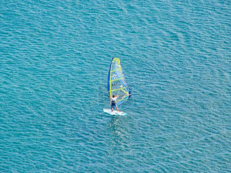 A windsurfer is sailing on his board with a sail on a calm azure sea water on a calm sunny day.