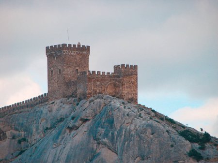 Eine antike mittelalterliche Festung am Rande einer Klippe ist von großen, hellen Wolken umgeben. Landschaft eines touristischen und historischen Ortes mit kaltem Blauton.