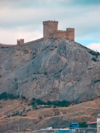 An ancient medieval fortress on the edge of a cliff is surrounded by large, bright clouds. Landscape of a tourist and historical place with a cold blue tint.