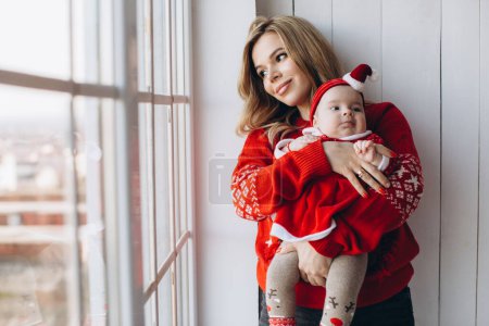 Téléchargez les photos : Portrait de mère et petite fille dans des vêtements de Noël traditionnels dans une pièce en bois clair près de la fenêtre, espace de copie - en image libre de droit