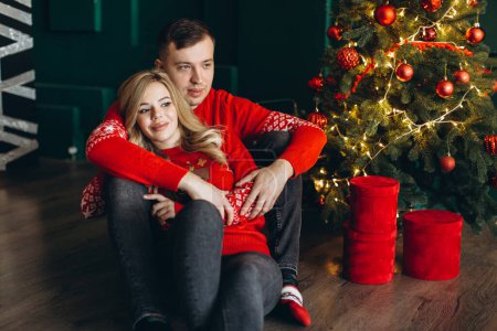 Photo for A charming couple in love in traditional Christmas red sweaters spend time near the Christmas tree - Royalty Free Image