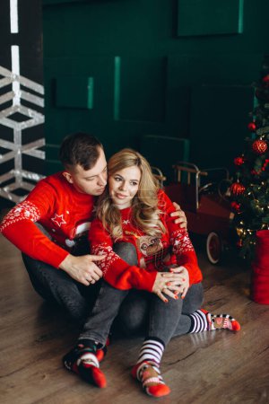 Téléchargez les photos : Un charmant couple amoureux en pull rouge de Noël traditionnel passe du temps près du sapin de Noël - en image libre de droit
