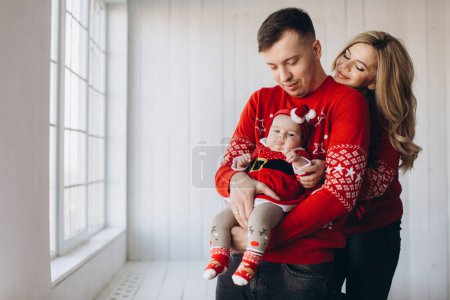 Foto de Retrato de la familia feliz mamá papá y su hija pequeña en rojo ropa de Navidad tradicional pasar tiempo juntos en la habitación de madera de luz cerca de la ventana, espacio de copia - Imagen libre de derechos
