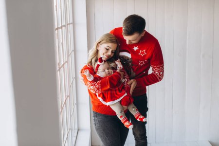 Photo for Happy family mom dad and little daughter in red traditional christmas outfit spending time together in light wooden room near window, copy space - Royalty Free Image