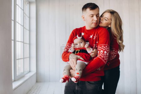 Téléchargez les photos : Portrait de maman de famille heureuse papa et petite fille en rouge vêtements de Noël traditionnels passer du temps ensemble dans la pièce en bois léger près de la fenêtre, espace de copie - en image libre de droit