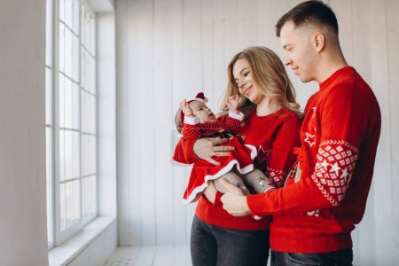 Téléchargez les photos : Portrait de maman de famille heureuse papa et petite fille en rouge vêtements de Noël traditionnels passer du temps ensemble dans la pièce en bois léger près de la fenêtre, espace de copie - en image libre de droit