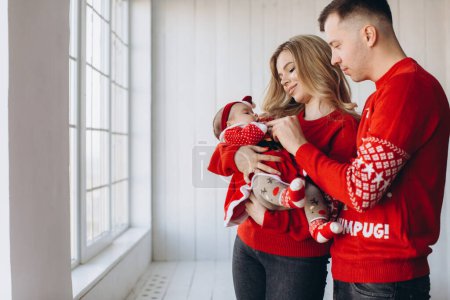 Photo for Portrait of happy family mom dad and little daughter in red traditional christmas clothes spending time together in light wooden room near window, copy space - Royalty Free Image