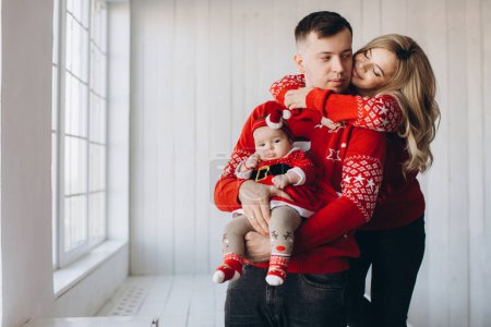 Photo for Portrait of happy family mom dad and little daughter in red traditional christmas clothes spending time together in light wooden room near window, copy space - Royalty Free Image