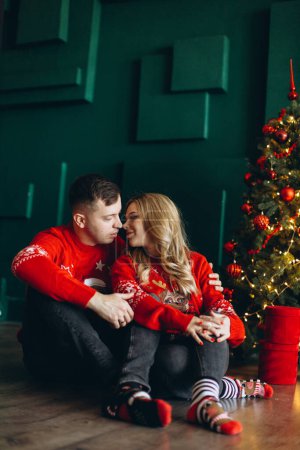 Téléchargez les photos : Un charmant couple amoureux en pull rouge de Noël traditionnel passe du temps près du sapin de Noël - en image libre de droit