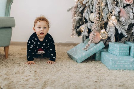 Téléchargez les photos : Un petit garçon bouclé dans un pull de Noël traditionnel passe du temps à la maison près du sapin de Noël - en image libre de droit
