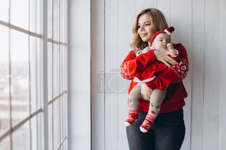 Photo for Portrait of mother and little daughter in traditional Christmas clothes in light wooden room near window, copy space - Royalty Free Image