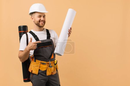 Photo for Positive construction engineer examines a drawing and holds a tube on a beige background - Royalty Free Image