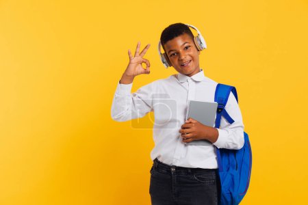 Photo for Happy African schoolboy wearing headphones and backpack holding books and notebooks and showing okay. Back to school concept. - Royalty Free Image