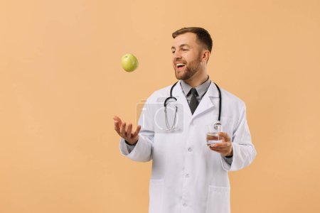 Foto de El médico nutricionista masculino con estetoscopio sonriendo y sosteniendo el agua y la manzana sobre fondo beige, concepto de plan de dieta - Imagen libre de derechos