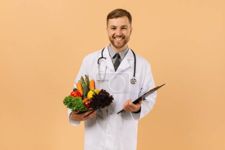 Foto de El médico nutricionista masculino con estetoscopio sosteniendo verduras frescas sobre fondo beige, concepto de plan de dieta - Imagen libre de derechos