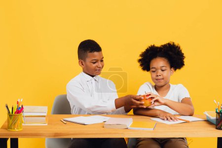 Happy african american schoolgirl and mixed race schoolboy sitting together at desk and sharing apple on yellow background. Back to school concept.