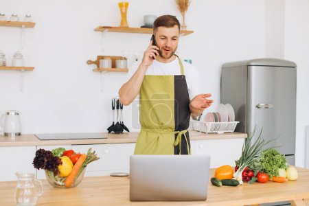 Photo for Attractive young man is cooking in the kitchen with a laptop on the table, talking on a smartphone. - Royalty Free Image