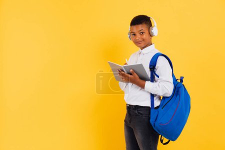 Téléchargez les photos : Heureux écolier africain portant des écouteurs et sac à dos tenant des livres et des cahiers. Concept de retour à l'école. - en image libre de droit