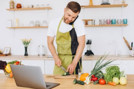 Photo for Attractive young man is cooking in the kitchen with a laptop on the table, talking on a smartphone. - Royalty Free Image