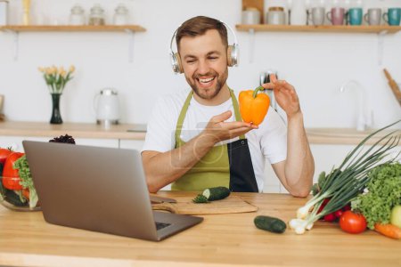 Foto de Hombre feliz en auriculares sentados en la mesa de la cocina y preparando ensalada, sosteniendo pimienta y mostrándola a la computadora portátil. - Imagen libre de derechos