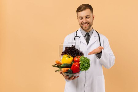 Foto de El nutricionista médico varón feliz con estetoscopio muestra verduras frescas en fondo beige, concepto de plan de dieta - Imagen libre de derechos