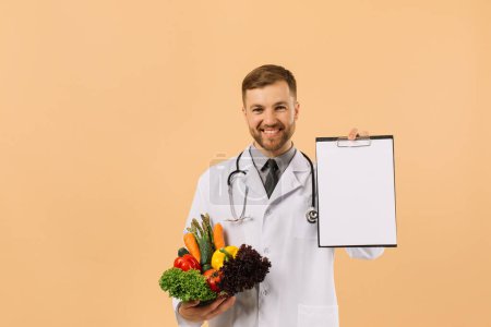 Foto de El médico nutricionista masculino con estetoscopio sosteniendo verduras frescas y carpeta con espacio de copia con sobre fondo beige - Imagen libre de derechos