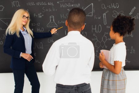 Photo for A female teacher assigns homework to school children near the blackboard. International team. Back to school. - Royalty Free Image