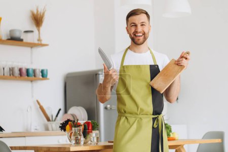 Téléchargez les photos : Portrait de l'homme heureux tenant conseil de cuisine et couteau dans la cuisine à la maison. - en image libre de droit