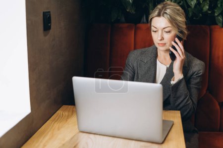 Téléchargez les photos : Femme d'affaires mature administrateur de restaurant assis parlant au téléphone et travaillant sur ordinateur portable - en image libre de droit