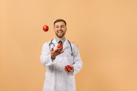 Foto de El médico nutricionista masculino con estetoscopio sonriendo y malabares tomates en fondo beige, concepto de plan de dieta - Imagen libre de derechos