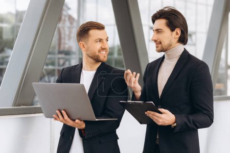 Photo for Two modern happy businessmen with laptop and document folder discussing something against the background of urban offices and buildings - Royalty Free Image
