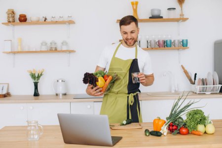 Photo for Portrait of happy man at home, man cooking vegetable salad looking at camera and smiling, slicing vegetables, using laptop for online cooking training - Royalty Free Image