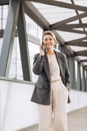 Téléchargez les photos : Portrait d'une belle femme d'affaires mature en costume et veste grise souriante et parlant au téléphone sur fond de bâtiments urbains et de bureaux modernes - en image libre de droit