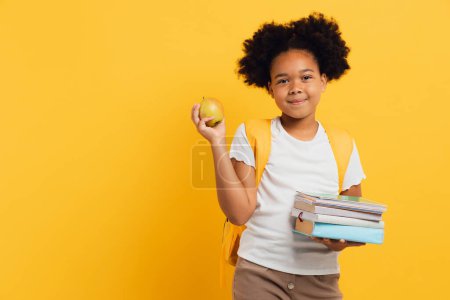 Téléchargez les photos : Heureuse écolière afro-américaine tenant des cahiers, des livres et un déjeuner pomme sur fond jaune, espace de copie. - en image libre de droit