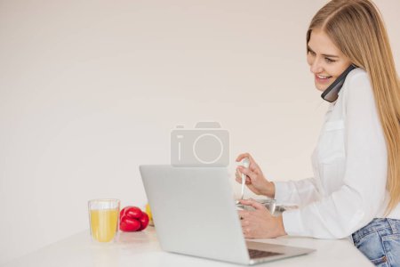 Téléchargez les photos : Femme blonde heureuse et souriante parlant au téléphone et à la recherche d'une recette sur Internet sur un ordinateur portable dans la cuisine - en image libre de droit