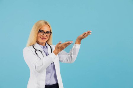 Photo for Mature woman doctor in glasses with stethoscope holding folder and show hand something on copy space the blue background - Royalty Free Image